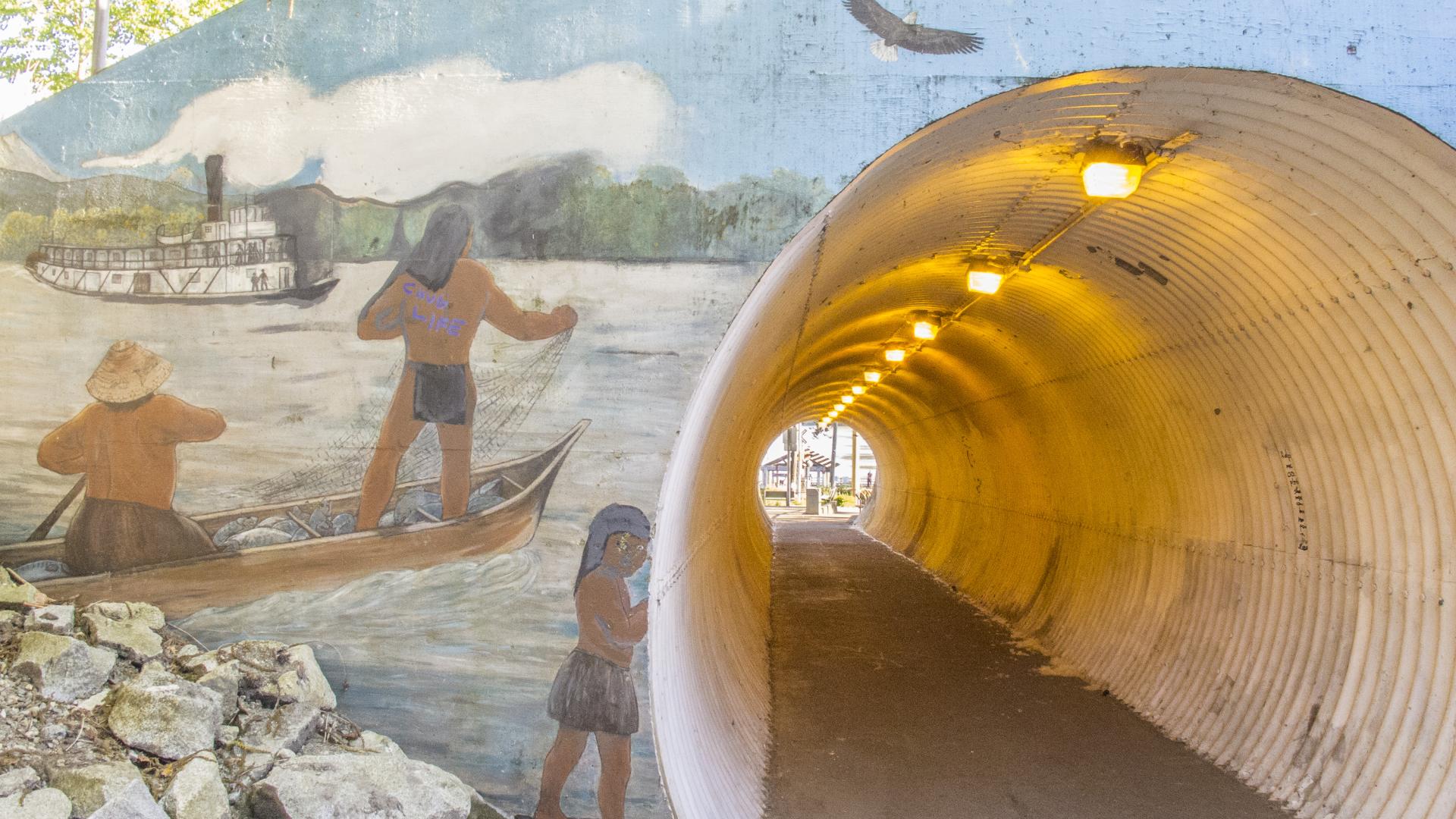 A mural depicting indigenous peoples fishing on the Fraser river in canoes covers the entrance to a tunnel along the Haney Heritage Walk. 